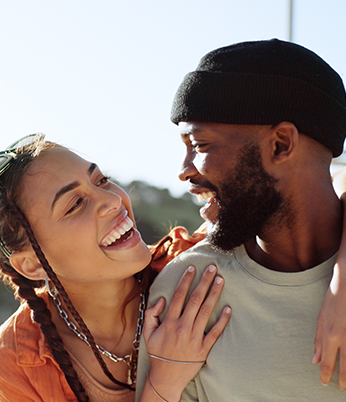  Um casal está sorrindo e olhando um para o outro de forma carinhosa; a mulher tem tranças e a pele clara, enquanto o homem tem barba e está usando um gorro preto.