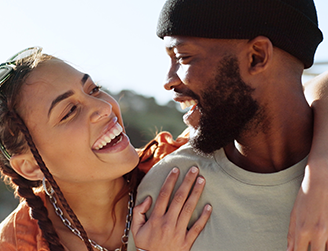  Um casal está sorrindo e olhando um para o outro de forma carinhosa; a mulher tem tranças e a pele clara, enquanto o homem tem barba e está usando um gorro preto.