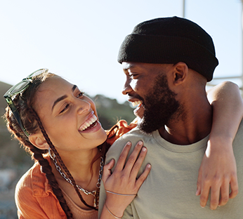  Um casal está sorrindo e olhando um para o outro de forma carinhosa; a mulher tem tranças e a pele clara, enquanto o homem tem barba e está usando um gorro preto.