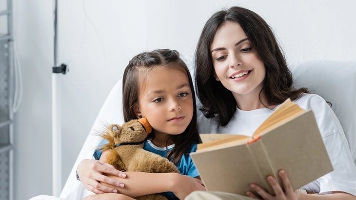 Uma mulher deitada em uma cama de hospital lê um livro para uma menina que está em seu colo, abraçando um ursinho de pelúcia.
