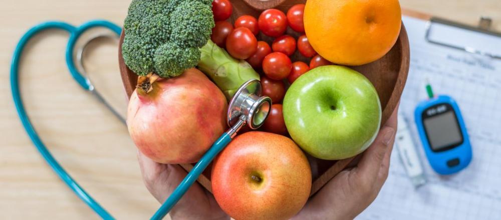 Pessoa segurando uma cesta de frutas e legumes, com um estetoscópio e um medidor de glicose ao fundo