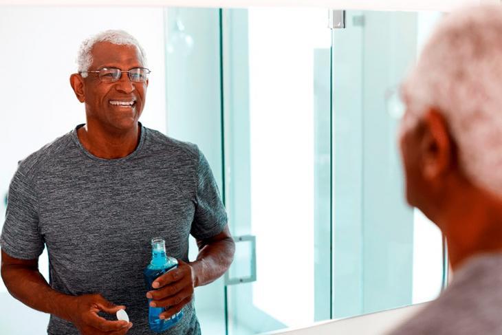 Imagem de um homem idoso segurando um frasco de enxaguante bucal enquanto se olha no espelho