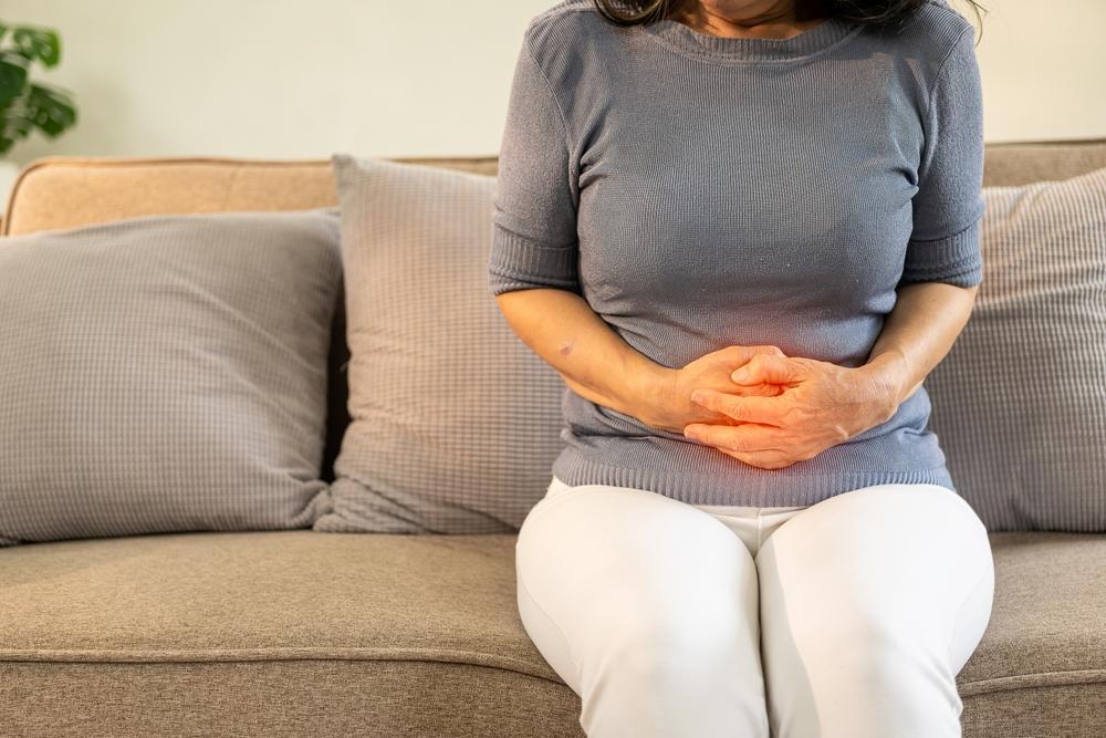 Mulher sentada no sofá segurando o abdômen com expressão de dor, representando sintomas digestivos, inflamação intestinal e a importância do diagnóstico precoce da doença inflamatória intestinal.