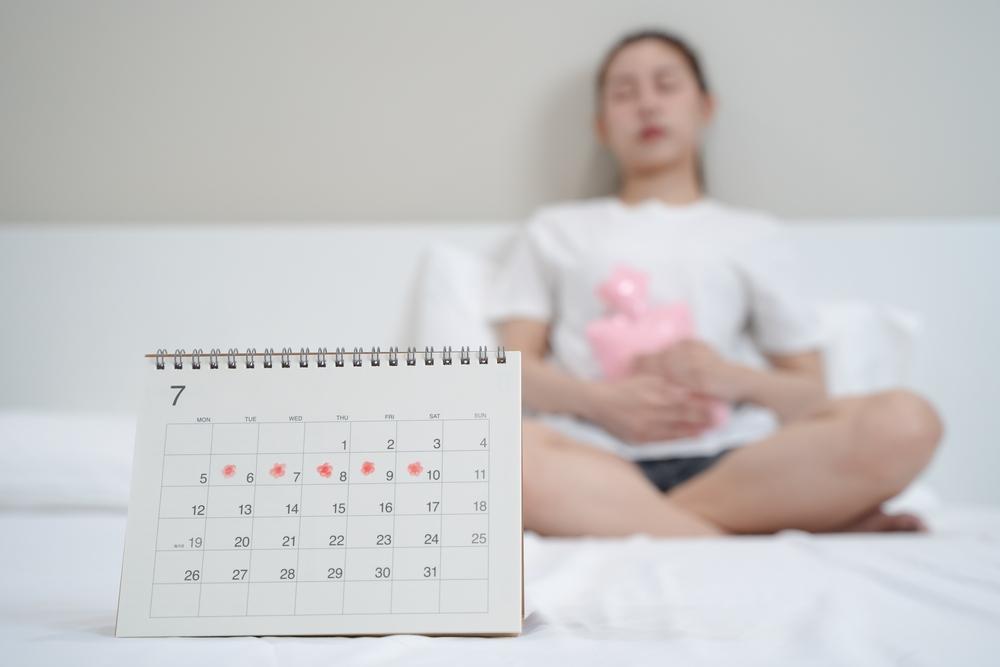 Calendário com marcações de ciclo menstrual em destaque e mulher ao fundo segurando o abdômen, representando fluxo menstrual irregular, sintomas hormonais e investigação além da TPM.