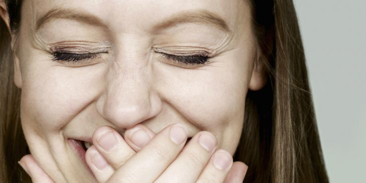 Close do rosto de uma mulher sorrindo e cobrindo parte da boca com as mãos