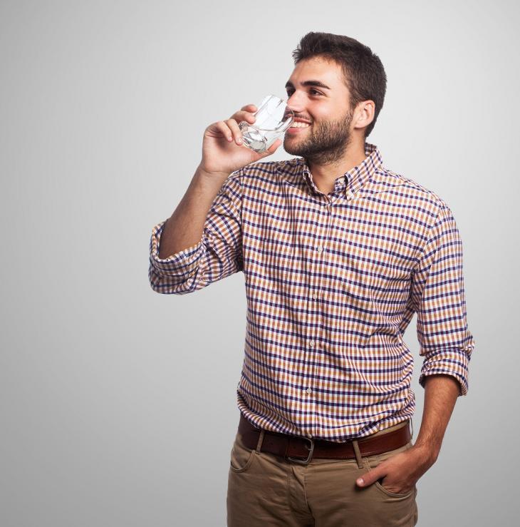 Imagem de homem bebendo água em um copo de vidro incolor, indicando o problema    de boca seca